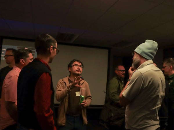 A group of men are engaged in a discussion in a dimly lit room, with one man in the center gesturing while holding a green can.