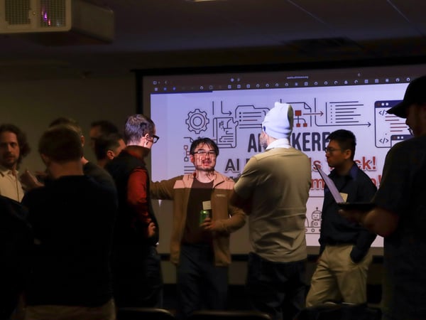 A group of people are gathered and talking at a tech event, with a large screen in the background displaying 'AI Tinkerers' content.