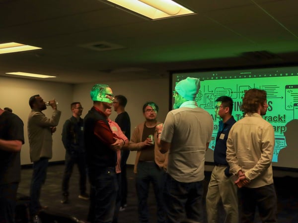 A group of people are gathered in a dimly lit room for a presentation or networking event, with a large green screen displaying text about an AI Tech Track.