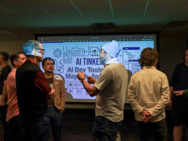 A group of men are gathered in a dimly lit room, engaged in conversation in front of a projection screen advertising an 'AI Tinker' event.