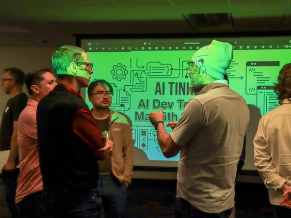 A group of people are gathered in a dimly lit room, engaged in a discussion in front of a large projection screen displaying 'AI TINKER'.