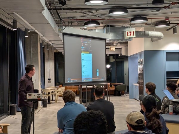 A man is giving a presentation to a seated audience in a modern office space, with a large projector screen showing a mobile app interface.