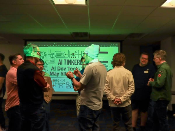 A group of people are gathered in a dimly lit room, standing and talking in front of a projector screen displaying an 'AI TINKERERS' event slide.