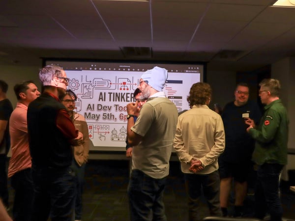 A group of people are gathered in a dimly lit room, engaged in conversation in front of a projection screen displaying an event announcement for 'AI Tinkerers'.
