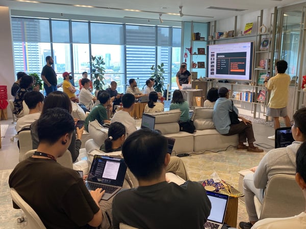 A group of people are gathered in a modern, well-lit office space for a tech presentation. A woman stands near a large screen displaying an agenda while the audience sits on lounge-style seating with laptops open.