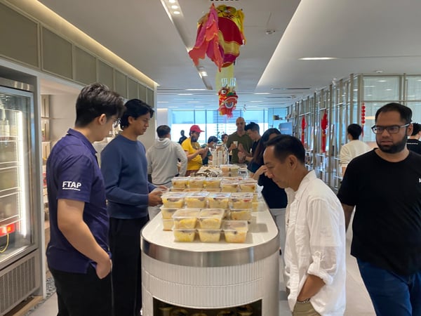 A group of people gathered in a modern indoor space, likely an office cafeteria, around a counter stacked with food containers, with festive decorations hanging from the ceiling.