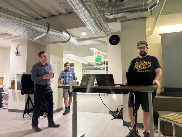 A photograph capturing three men in a modern office space with exposed ceiling ductwork, where one man is giving a presentation while another operates a laptop at a standing desk.