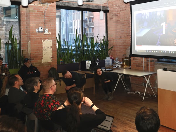 A group of people are gathered in a brick-walled room for a presentation or demonstration. A woman stands near a projection screen displaying a large number '1' and skeletal graphics.