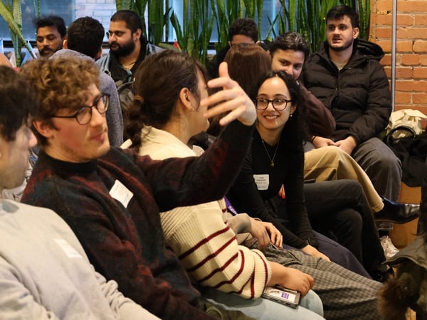 A group of people are gathered indoors, likely for a workshop or social event, with one person gesturing while others listen and converse.