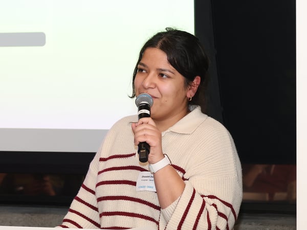 A woman is speaking into a microphone during a presentation, wearing a striped sweater and a name tag.
