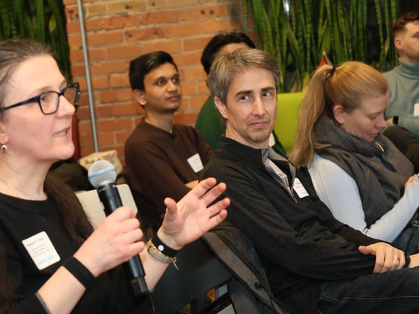 A woman with glasses speaks into a microphone during an indoor event while others in the background listen.