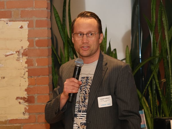 A man wearing a pinstripe blazer and glasses is speaking into a handheld microphone at an indoor event. He has a name tag and is standing in front of a brick wall and green plants.