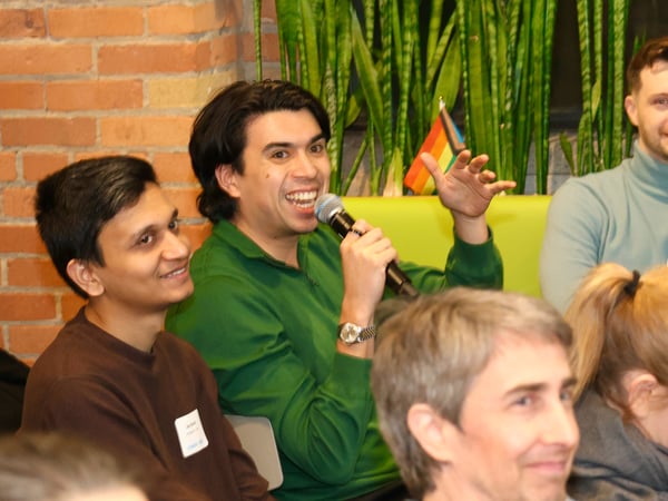 A man in a green sweater enthusiastically speaks into a microphone during an indoor event, while others listen nearby against a backdrop of a brick wall and plants.