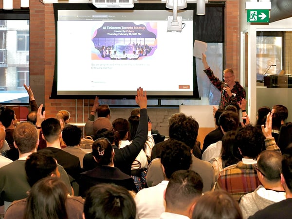 A speaker addresses a crowd at an 'AI Tinkerers Toronto Meetup' event, with several audience members raising their hands in front of a large projection screen.