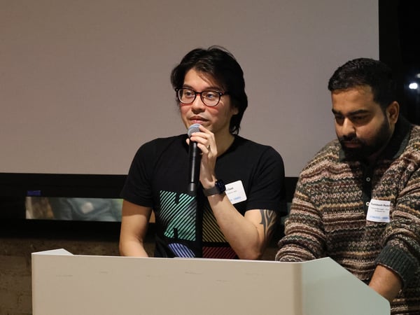 Two men stand at a podium during an indoor event; one man speaks into a microphone while the other looks down.