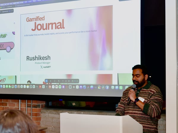 A man stands at a white podium giving a presentation, with a large screen behind him displaying a slide for a 'Gamified Journal' project.