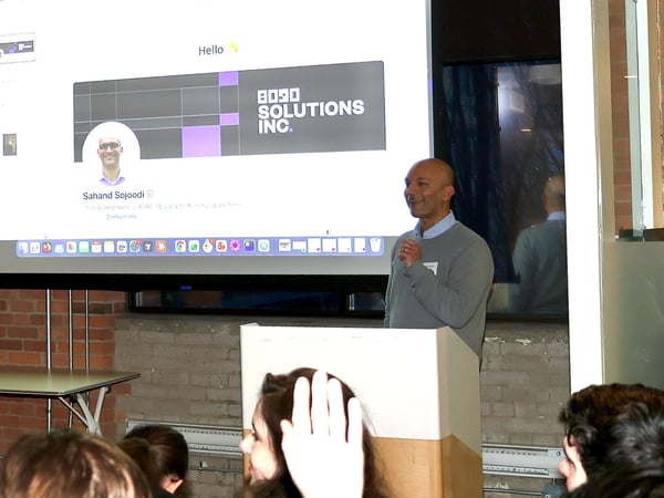 A man is giving a presentation at a podium in front of a large screen displaying a LinkedIn profile for Sahand Sojoodi and the logo for 8090 SOLUTIONS INC.
