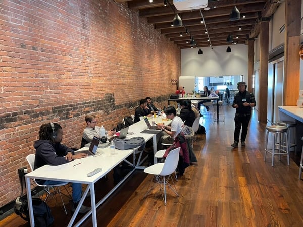 A group of people are working on laptops in a modern, industrial-style coworking space featuring an exposed brick wall and wooden floors.