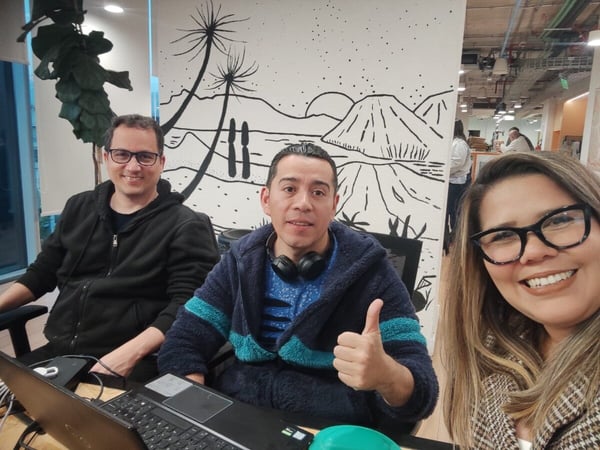 Three colleagues, two men and one woman, are smiling and posing for a photo in an office environment with a large black-and-white mural in the background.