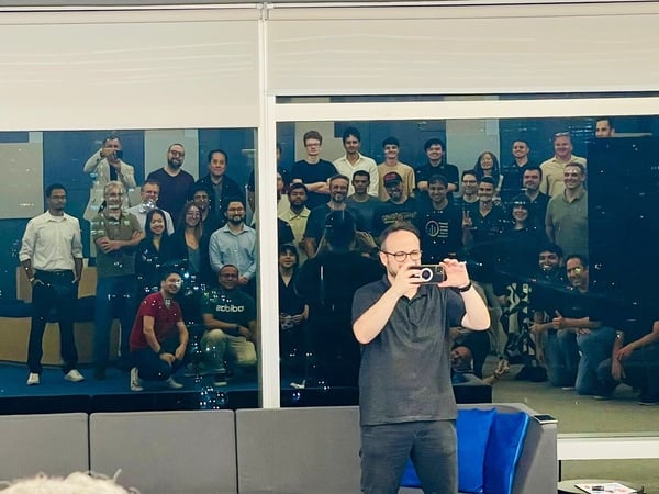 A group of approximately 35 people is posing for a photo behind a large glass partition in a modern office or event space, while a man in the foreground takes a picture of the group using a smartphone.
