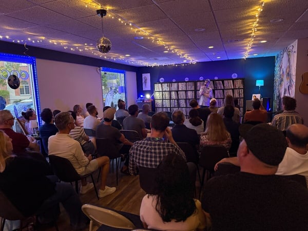 An indoor photograph of an audience seated in chairs, watching a performer stand at the front of a cozy room decorated with string lights and shelves of vinyl records.