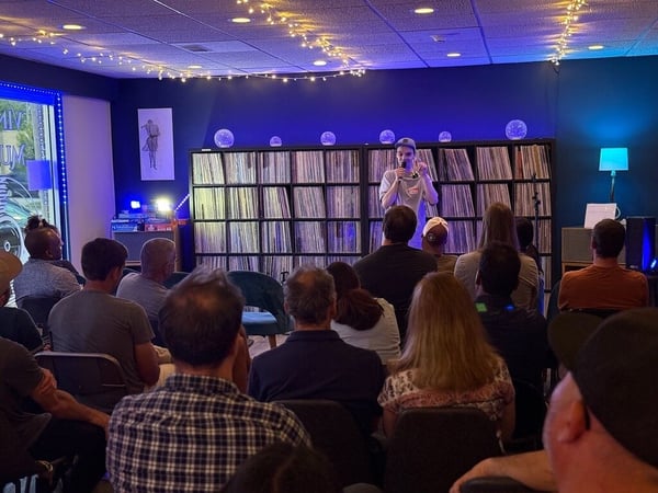 A young man stands on a small stage speaking into a microphone in front of an audience seated in a dimly lit room with shelves of vinyl records in the background.