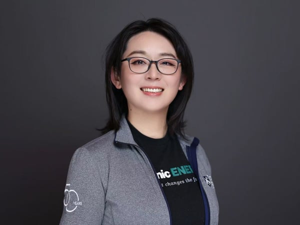 A professional headshot of a smiling woman wearing glasses, a gray zip-up jacket, and a black t-shirt with corporate branding, set against a solid dark gray background.