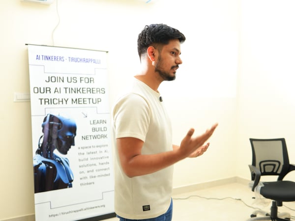 A young man in profile is gesturing while speaking at an event, with an 'AI Tinkerers' meetup banner in the background.