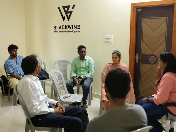 A group of people are seated in a circle for a discussion inside an office room that features a company logo on the wall.