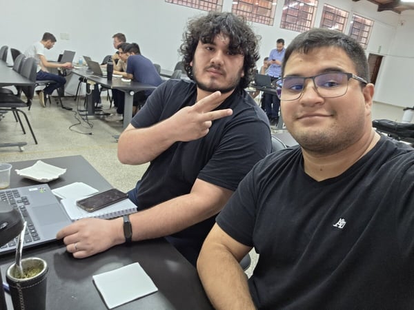 Two young men are posing for a photo at a desk in a room filled with other people working on laptops.