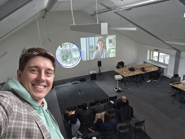 A man takes a selfie in a large, modern event space where an audience is watching a video presentation projected onto a wall.