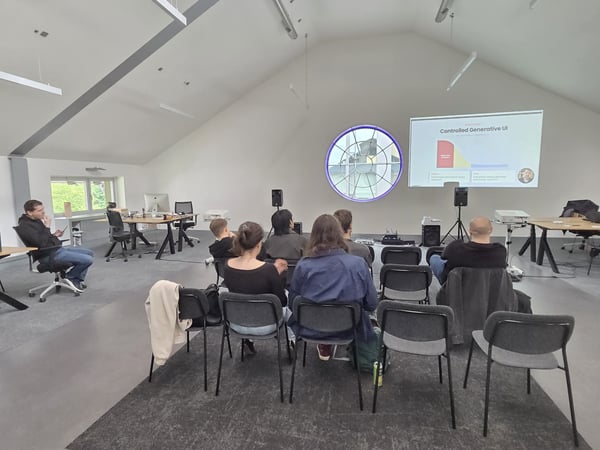A group of people are seated in a large, minimalist white room with a high ceiling, watching a presentation about 'Controlled Generative UI' projected onto a wall.