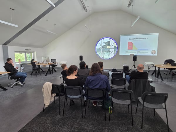 A group of people are seated in a large, modern room with vaulted ceilings, watching a presentation titled 'Controlled Generative UI' projected onto a wall.