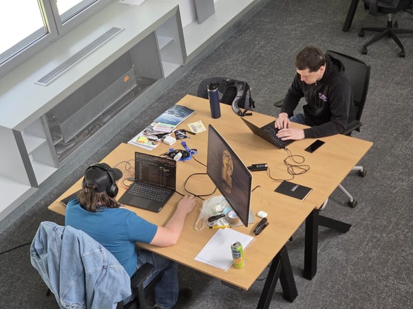 Two men are working on their laptops at a large wooden desk in a modern office environment.
