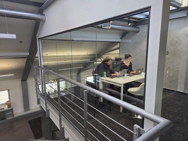 Two individuals are seen working on their laptops at a white table inside a glass-walled office space with modern industrial architecture.