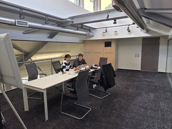 Two men are sitting at a long white table in a modern, well-lit office or meeting space, working on their laptops.
