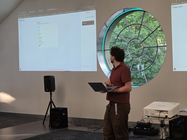 A man stands in a modern room holding a laptop, presenting a software interface projected onto a large white wall next to a distinctive circular window.