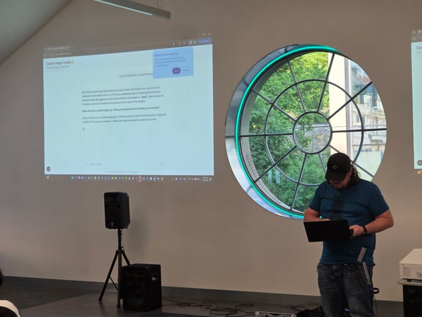 A man stands in a room holding a laptop in front of a projector screen and a large circular window. The screen displays a website titled 'Coder's Magic Cookie' with humorous text about debugging a server.