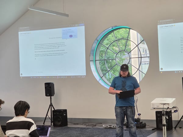 A man in a blue shirt and black cap is giving a presentation using a laptop in front of two large projected screens in an indoor space.