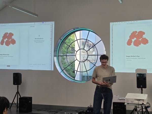 A man stands in a room holding a laptop, positioned between two projector screens displaying a 'Happy Mother's Day' message. A large circular window is centered behind him, showing a view of trees and buildings outside.