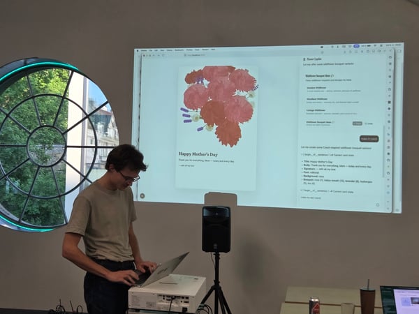A man stands at a desk using a laptop, demonstrating a software interface projected onto the wall behind him that shows a Mother's Day card design tool.