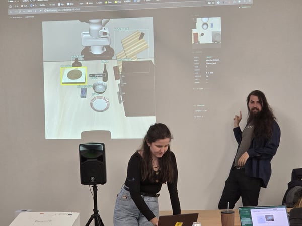 A man and a woman are giving a technical presentation or demonstration, with a projection showing a 3D robotic simulation on the wall behind them.