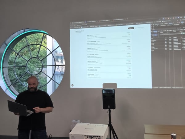 A man holding a laptop stands in front of a projected screen displaying a web application and developer tools during a presentation.