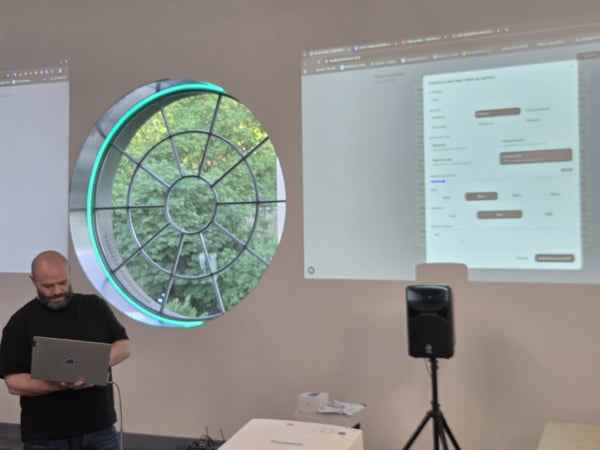 A man stands with a laptop in front of a projection showing a software interface, next to a circular window with green lighting.