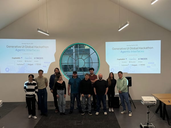A group of ten people posing for a photo in an indoor event space with two projection screens displaying hackathon information and a circular window in the background.