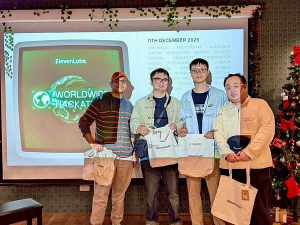 Four men stand side-by-side in an indoor event space, posing in front of a projection screen advertising the 'ElevenLabs WORLDWIDE HACKATHON.' The men are holding branded tote bags, and the room is decorated with Christmas garland and a Christmas tree.