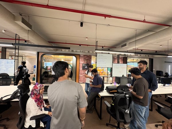 A group of young professionals are gathered in a modern, industrial-style office space, engaged in conversation and looking at screens or whiteboards.