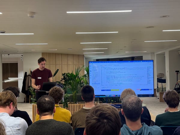 A man in a maroon shirt is giving a presentation to an audience, with a large screen behind him displaying lines of Python code.