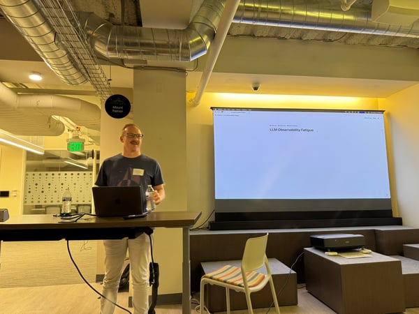 A man is giving a presentation in a modern office space, standing behind a desk with a laptop while a large projection screen displays the title "LLM Observability Fatigue."