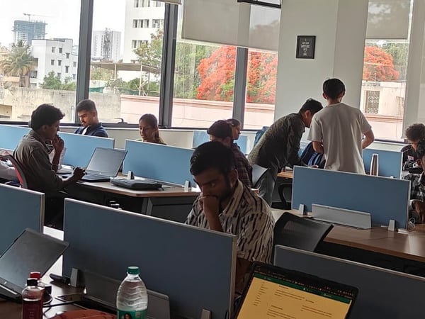 A photograph of a busy office environment where several people are working at desks equipped with laptops and blue dividers.
